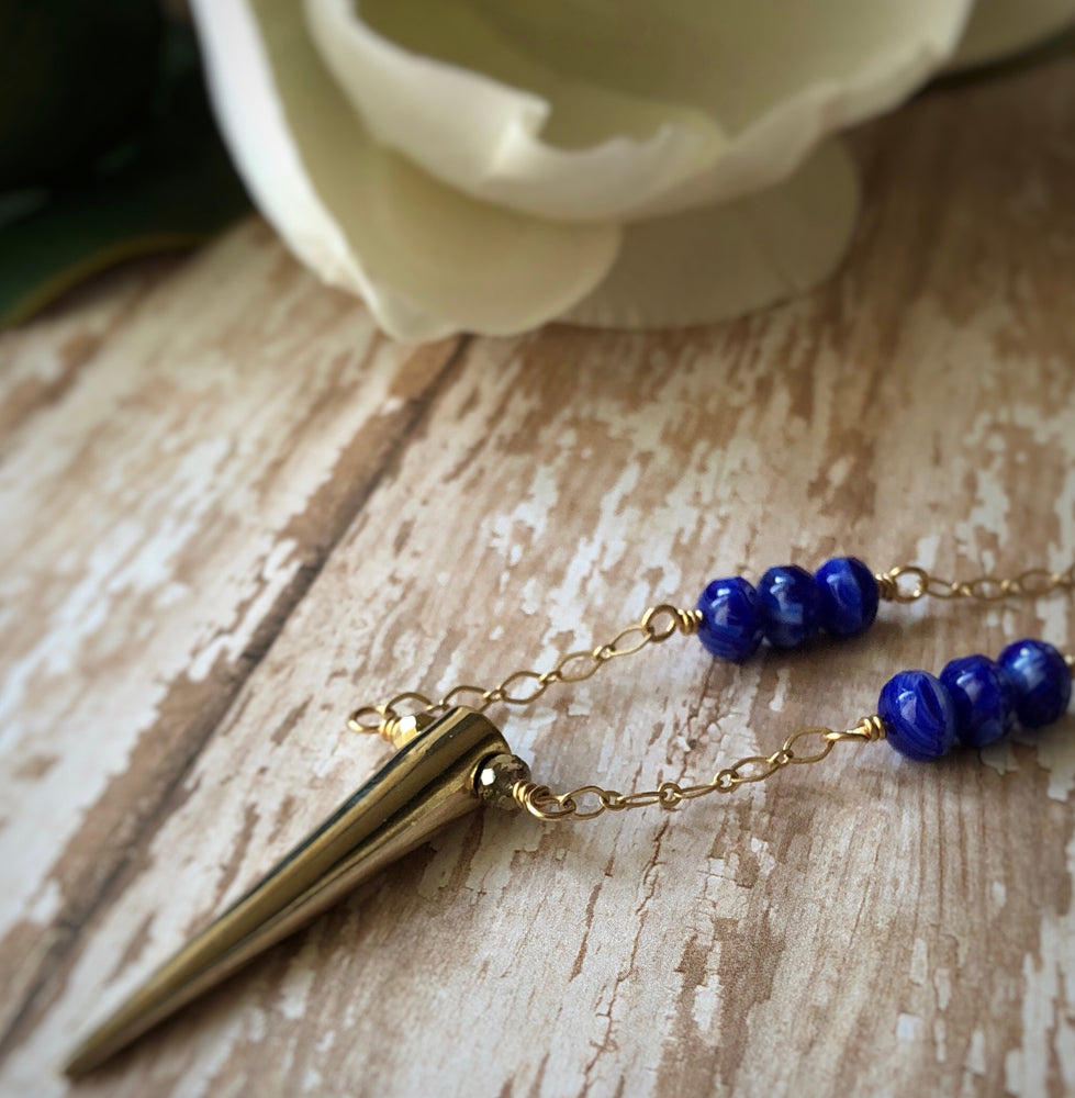 A gold-toned spike-pendant necklace with blue beads lies on a wood-pattern background, with part of a flower visible a few inches away.