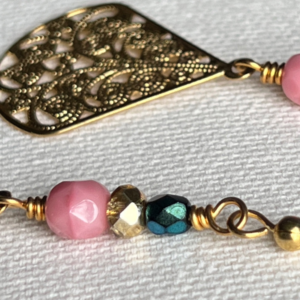 This photo features a close-up of the golden focal of one earring and the teal, golden, and pink beads of the other, all on an ivory fabric background.