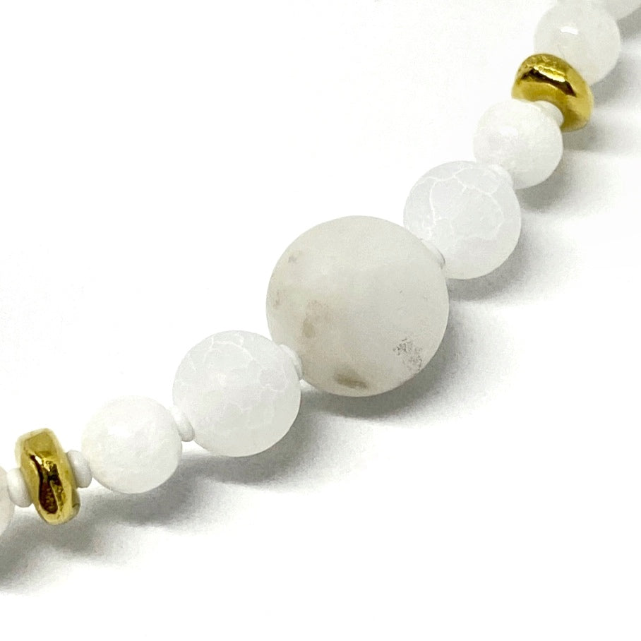 A strand of gold-toned metal spacer beads, round white beads, and tiny white spacer beads stretches from the lower left corner to the upper right, on a white background. The largest bead, in the center, has swirls of gray.