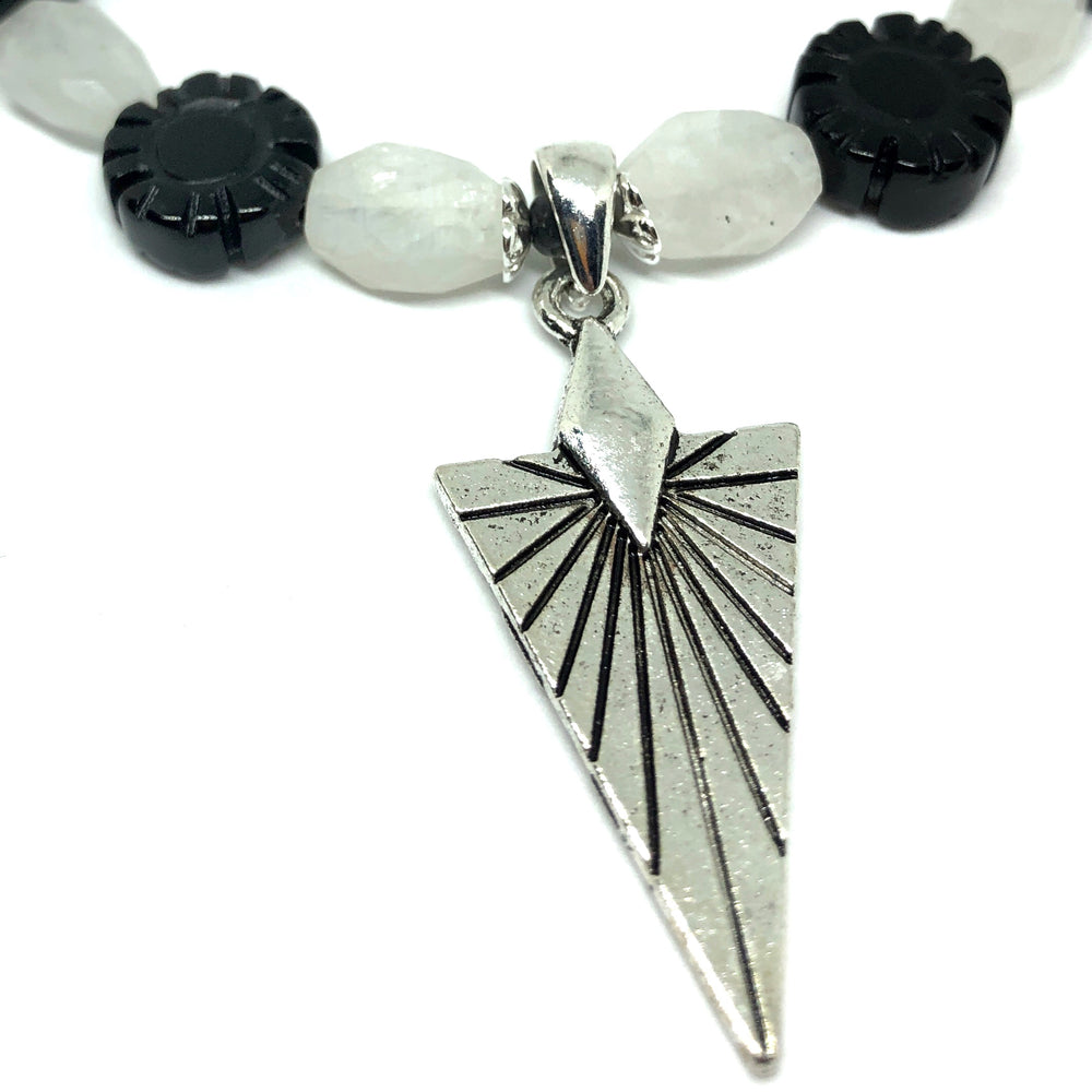 A line of black and white beads curves across the top part of the photo, and a silver-toned arrowhead-shaped pendant sits in the center. The pendant and beads are pictured at a slight angle and lie on a white background.