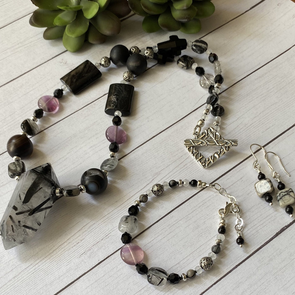 On a white wood-look background lie a pendant necklace, bracelet, and pair of earrings featuring black beads and silver-toned metals.