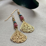 On an ivory fabric background lie two earrings, with their golden teardrop-shaped focals in the foreground and their teal, golden, and pink beads farther away. At the top of the photo a green leaf is partly visible.
