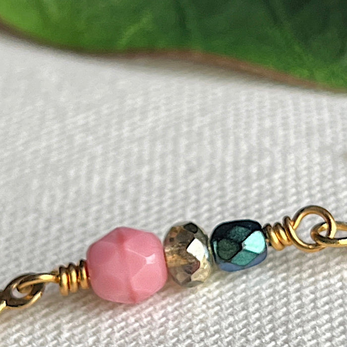 In the foreground of the photo is a piece of wire with wrapped loops at both ends, and on the wire is a larger pink bead and two smaller beads, one teal and one golden. In the background a green leaf is partly visible. The backdrop for the photo is ivory fabric.