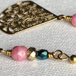 This photo features a close-up of the golden focal of one earring and the teal, golden, and pink beads of the other, all on an ivory fabric background.