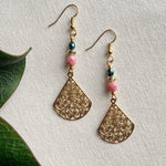 On an ivory fabric background lie two earrings featuring golden earwires and teardrop-shaped focals as well as teal, golden, and pink beads. To the left, two green leaves are partly visible.