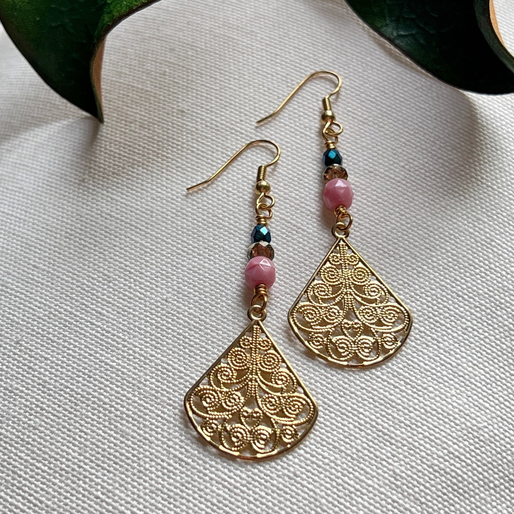 In the foreground of the photo is a piece of wire with wrapped loops at both ends, and on the wire is a larger pink bead and two smaller beads, one teal and one golden. In the background a green leaf is partly visible. The backdrop for the photo is ivory fabric.