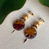 Pair of earrings with circular glass beads, crystal pearls, wood beads, and gold accents on a textured surface, with leaves at the top of the photo.