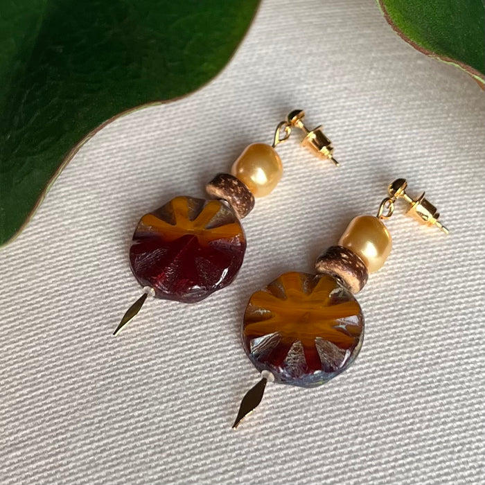 Pair of earrings with circular glass beads, crystal pearls, wood beads, and gold accents on a textured surface, with leaves at the top of the photo.