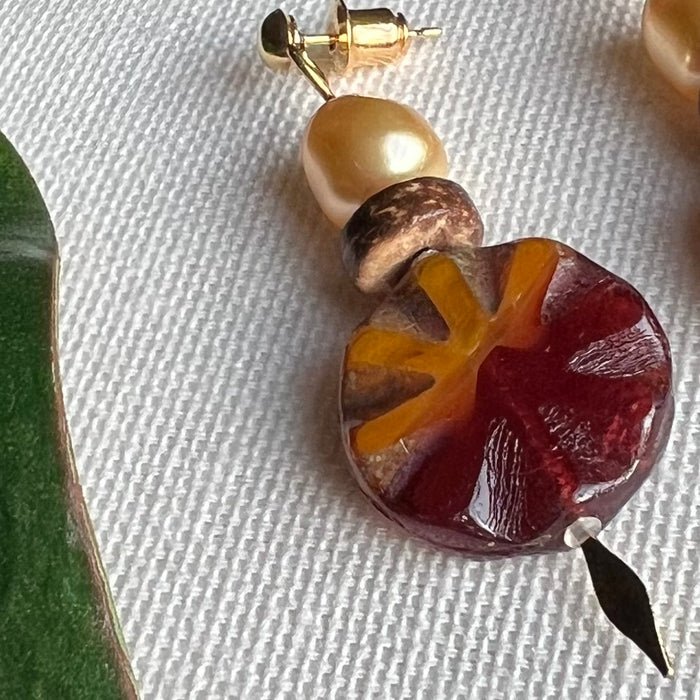 Earring with a crystal pearl, a wood bead, and maroon and orange Czech glass on an ivory background, with a green leaf to the left.