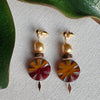 Pair of earrings with golden accents, crystal pearls, wood beads, and colorful circular designs on a textured surface.