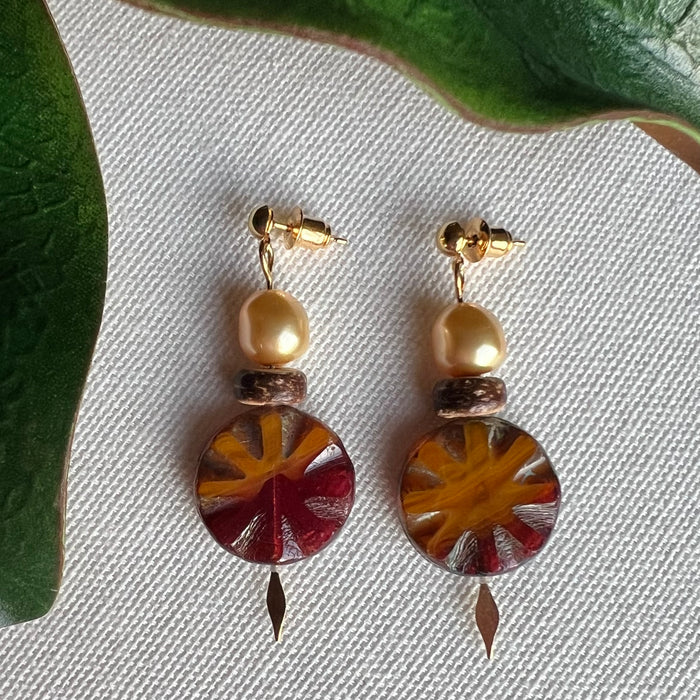 Pair of earrings with golden accents, crystal pearls, wood beads, and colorful circular designs on a textured surface.