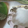 This photo features a close-up of a beaded bracelet with a silver butterfly-shaped clasp. In the foreground are warm-gray round beads alternating with light-pink butterfly-shaped ones. The bracelet lies on ivory fabric, framed by two green leaves.