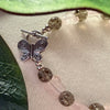 A beaded bracelet featuring a silver butterfly toggle clasp, warm-gray crackle-effect rounds, and light-pink butterfly-shaped beads lies on an ivory fabric, with green leaves to the top and side.