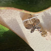 A silver butterfly toggle clasp lies on an ivory fabric background with green leaves above and below. To one side, a warm gray bead and a light pink one are visible.