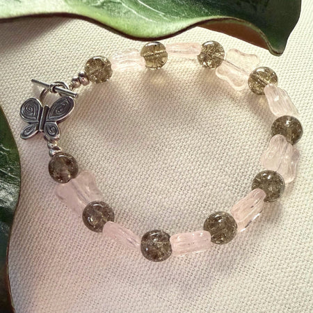 A beaded bracelet with light-pink butterfly shapes alternating with warm-gray crackle-effect rounds rests on an ivory fabric with green leaves to the top and side. The silver toggle clasp on the bracelet is butterfly-shaped.