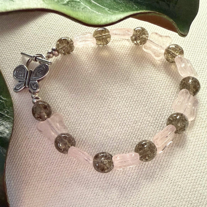 A beaded bracelet with light-pink butterfly shapes alternating with warm-gray crackle-effect rounds rests on an ivory fabric with green leaves to the top and side. The silver toggle clasp on the bracelet is butterfly-shaped.