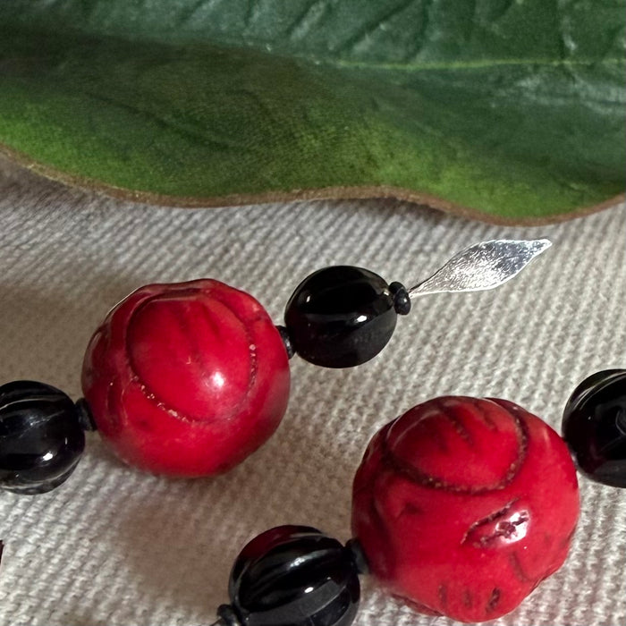 This photo is a close-up of small, fluted black beads to either side of larger, red carved beads. A silver-toned, spearhead-shaped tip extends to one side. The beads lie on textured ivory fabric with a green leaf in the background.