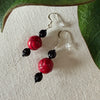 A pair of silver-toned earrings with red and black beads lies on a textured ivory fabric background, framed by green leaves along the top and side.