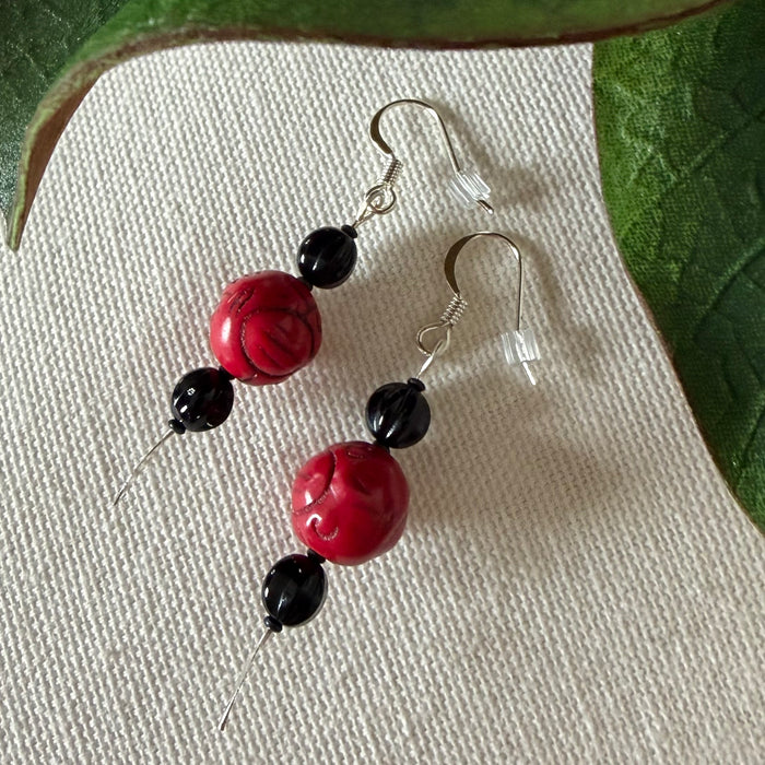 A pair of silver-toned earrings with red and black beads lies on a textured ivory fabric background, framed by green leaves along the top and side.