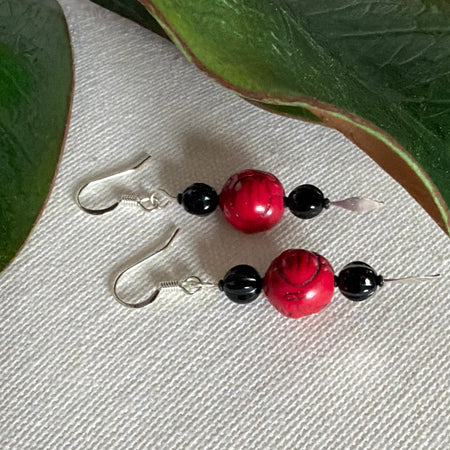A pair of silver-toned earrings with red and black beads lies on an ivory fabric background with green leaves to the top and sides. The black beads are fluted, and the red ones have a design carved into them.