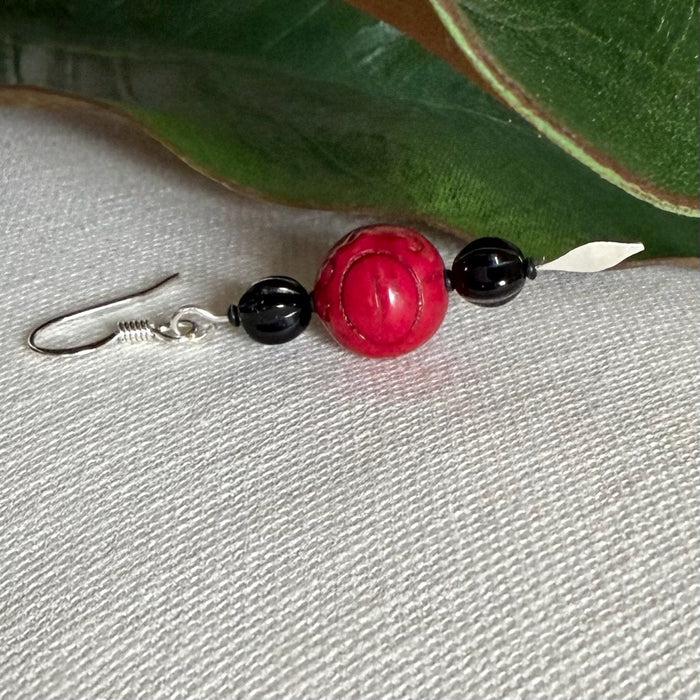 A single silver-toned earring with small, fluted black beads above and below a larger, carved red bead lies on a light-colored fabric with green leaves in the background. The bottom tip of the earring is shaped like a spearhead.