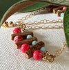 Green leaves frame two partly visible earrings with red and blue stone beads, brown wood beads, and gold chain on an ivory fabric background.