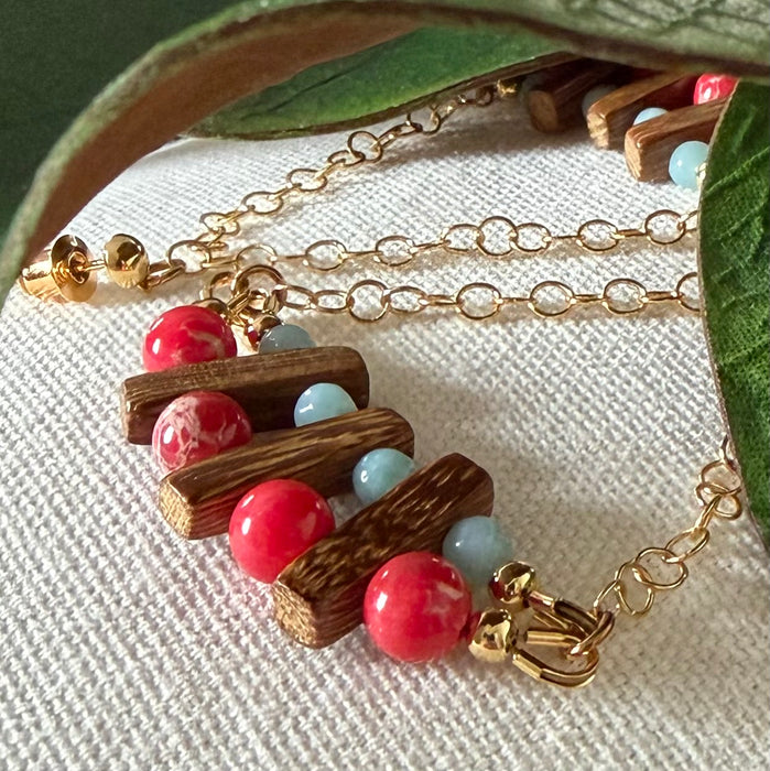 Green leaves frame two partly visible earrings with red and blue stone beads, brown wood beads, and gold chain on an ivory fabric background.