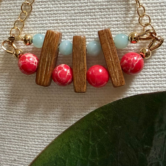 Two rows of beads, red and blue, connected by brown wood beads and suspended from gold-toned chain, lie on an ivory fabric background, with a green leaf in one corner.