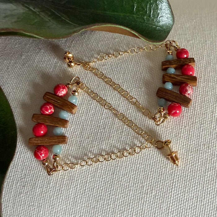 Large swing earrings with brown wood and red and blue stone beads suspended from gold-toned chain lie on an ivory textured background with green leaves along the side and top.