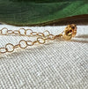 A gold-toned earring post with gold-toned chain attached lies on a textured ivory fabric background, in front of a green leaf.