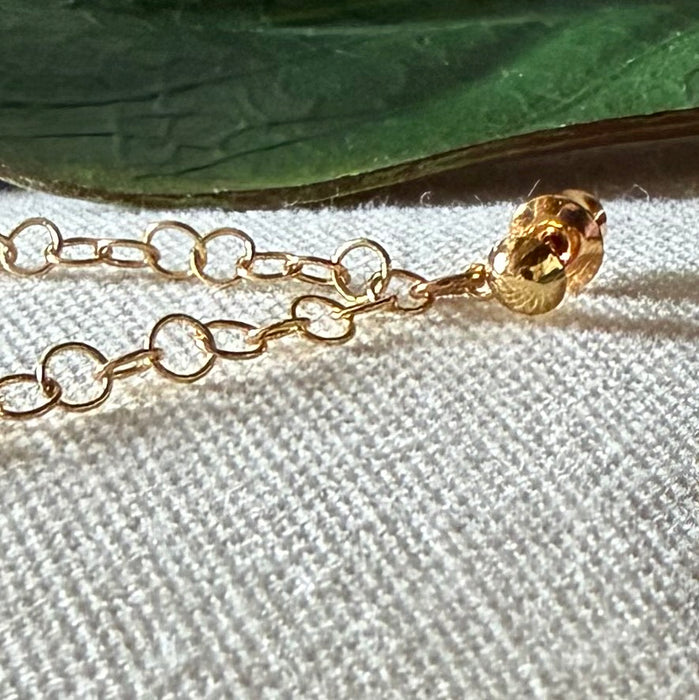 A gold-toned earring post with gold-toned chain attached lies on a textured ivory fabric background, in front of a green leaf.