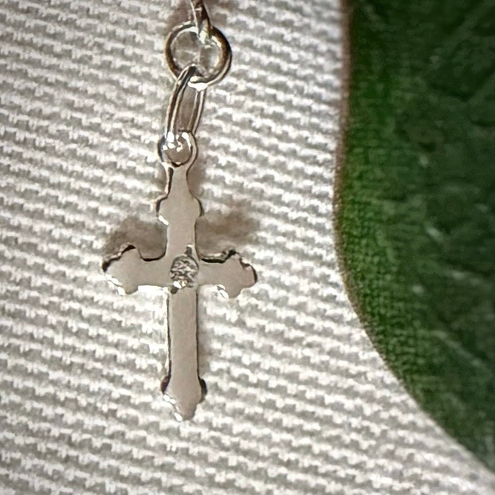 A silver cross charm, set with a small, clear stone, rests on a textured ivory surface with a green leaf to one side.