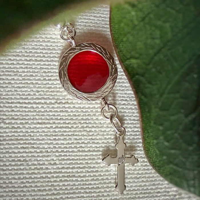 A silver dangle with a silver-framed red circle and a cross charm set with a small, clear stone rest on a textured ivory fabric background. Green leaves appear in one corner and along one side of the photo.