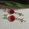 Silver post earrings with red circles,framed in silver, and cross charms featuring small, clear stones lie on a textured ivory fabric background. A green leaf appears in the top portion of the photo.