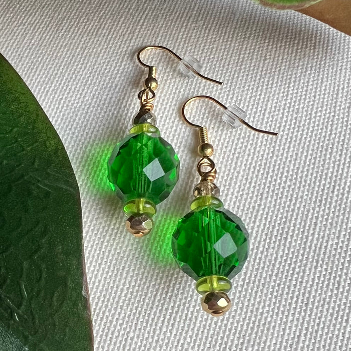 Angled view of green beaded earrings on a textured ivory fabric background with a green leaf to the left