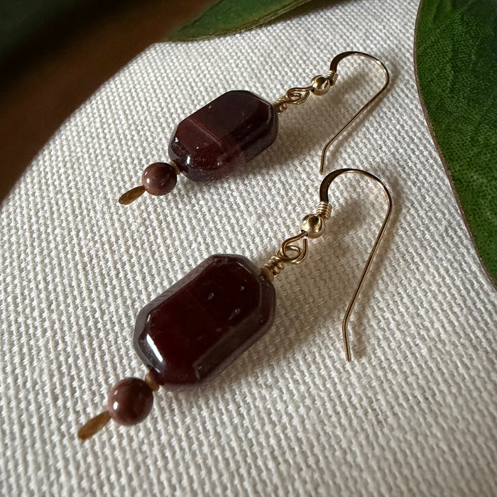 A pair of gold-toned earrings with dark, faceted, oblong octagonal beads accented with smaller round brown beads lies on ivory textured fabric, framed by green leaves at the top and side.