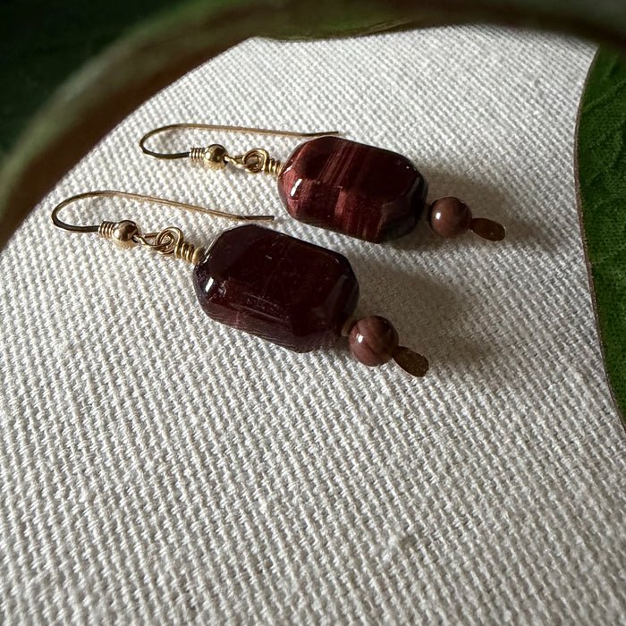A pair of gold-toned earrings with faceted dark burgundy beads in an oblong octagonal shape, accented with small brown round beads, lies on ivory fabric, framed by green leaves at the top and sides.