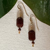 A pair of gold-toned earrings with faceted dark beads and smaller round brown beads rests on a textured ivory-colored fabric with green leaves in the top corners.