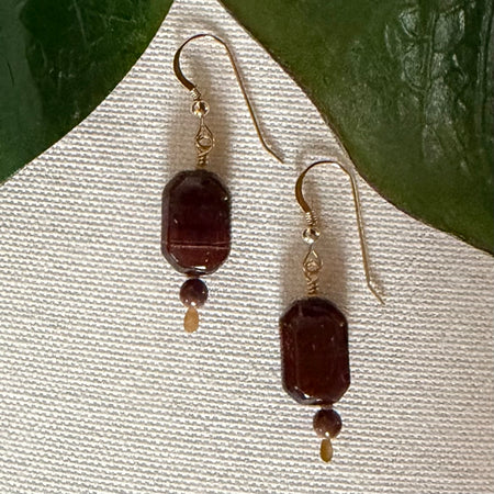 A pair of gold-toned earrings with faceted dark beads and smaller round brown beads rests on a textured ivory-colored fabric with green leaves in the top corners.