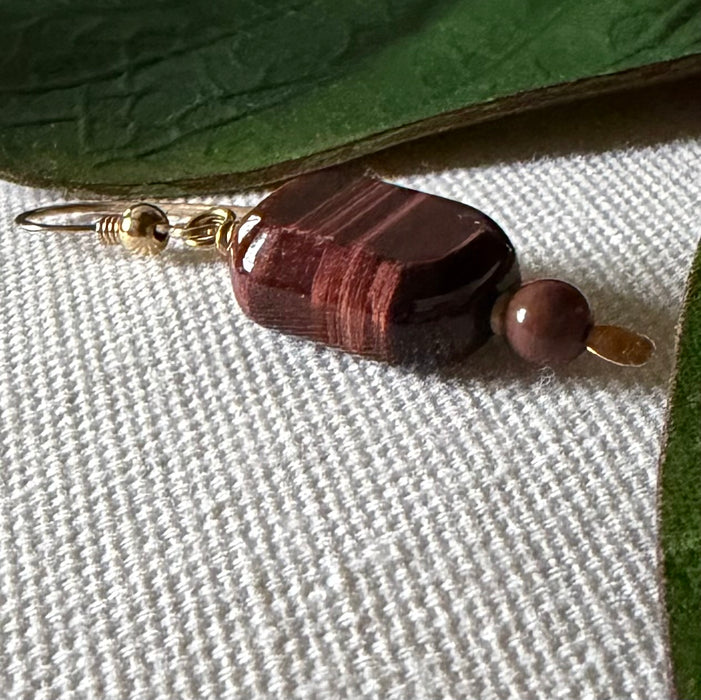 A gold-toned earring with a deep-burgundy, oblong-octagonal bead and a smaller, round brown bead lies on ivory fabric, framed on two sides by green leaves.
