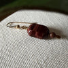 A single gold-toned earring featuring a deep-burgundy, oblong-octagonal bead accented with a smaller, round brown bead lies on an ivory fabric, with a blurred green leaf at the top of the photo.