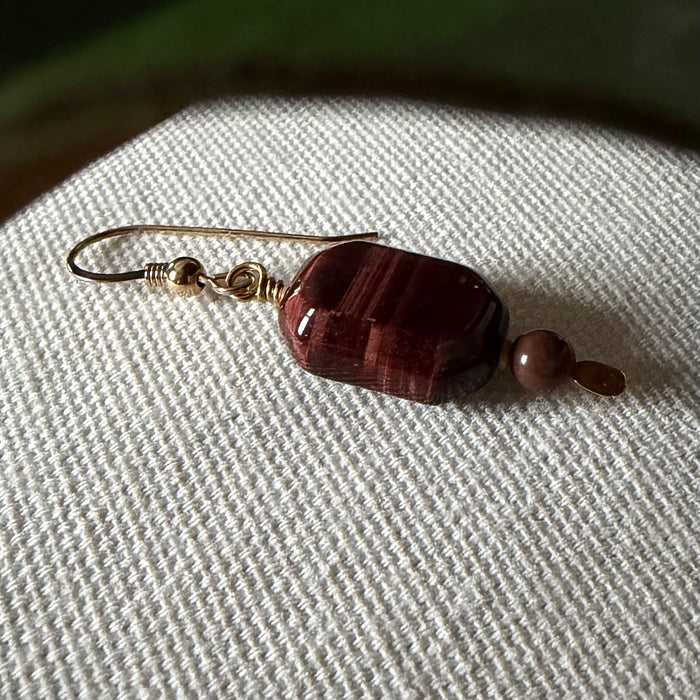 A single gold-toned earring featuring a deep-burgundy, oblong-octagonal bead accented with a smaller, round brown bead lies on an ivory fabric, with a blurred green leaf at the top of the photo.