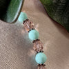 Part of a strand of opaque mint-green and transparent blush-pink beads, separated by thin silver spacers, rests on a neutral textured fabric, with green leaves at the top corners of the photo.