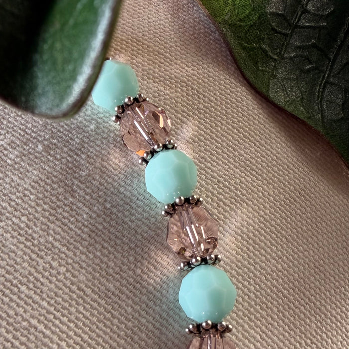 Part of a strand of opaque mint-green and transparent blush-pink beads, separated by thin silver spacers, rests on a neutral textured fabric, with green leaves at the top corners of the photo.