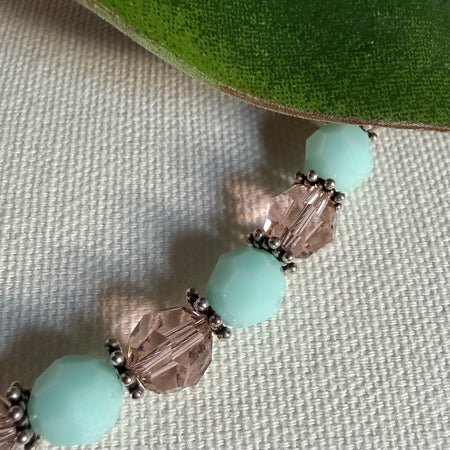 Part of a strand of opaque mint-green and transparent blush-pink beads, separated by thin silver spacer beads, lies on ivory textured fabric, with a green leaf at the top of the photo.