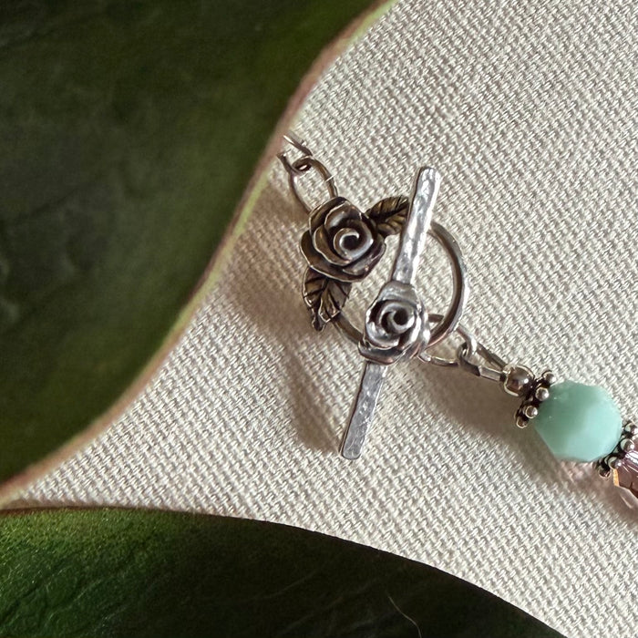 A silver toggle clasp, featuring a three-dimensional rose on the bar and a rose and leaves on the round, lies on textured ivory fabric, with green leaves to the left. A mint-green bead and silver spacers and findings are to the right of the clasp.