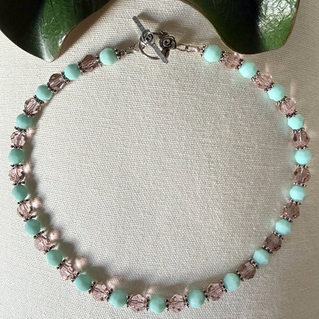 A necklace with opaque mint-green and transparent blush-pink beads lies on textured ivory fabric, with green leaves near the top corners of the photo.