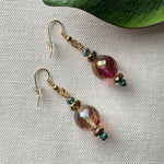 A pair of earrings featuring gold-toned earwires and green, golden, and blush-pink faceted beads lie on ivory fabric, with a green leaf partly visible in the upper right of the photo.