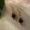 A pair of gold-toned earrings featuring pink, golden, and purple beads lies on ivory fabric. To the left is part of a green leaf, and in the top right is part of a pale pink translucent object.