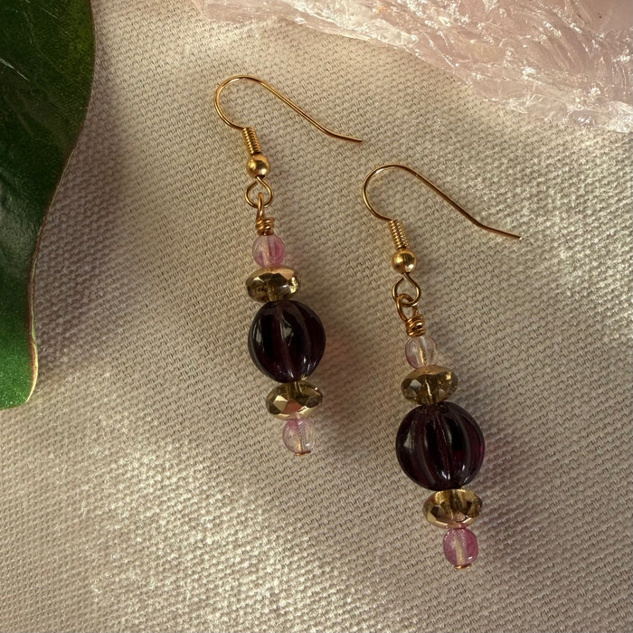 A pair of gold-toned earrings featuring pink, golden, and purple beads lies on ivory fabric. To the left is part of a green leaf, and in the top right is part of a pale pink translucent object.